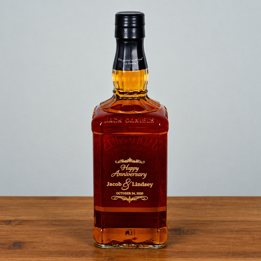A customized Jack Daniel's 16 oz square glass whiskey bottle with amber liquid, featuring a black cap and a personalized 'Happy Anniversary Jacob & Lindsey' label, set on a wooden surface against a neutral gray background.
