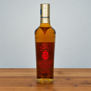 A bottle of macallan 12 with amber liquid, a red label, and a gold cap, standing on a wooden surface against a light gray background.