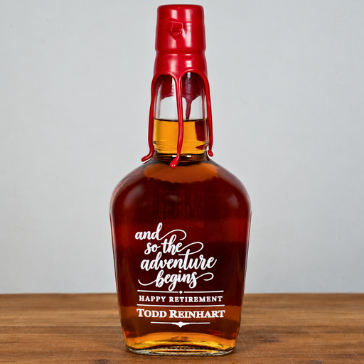 A custom engraved whiskey bottle with amber liquid, a red wax seal, and white text reading 'and so the adventure begins HAPPY RETIREMENT Todd Reinhart', set on a wooden surface against a light gray background.
