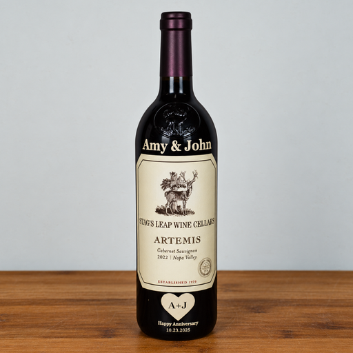 A custom dark glass wine bottle with a cream-colored label, standing on a wooden surface against a light gray background. The label features vintage-style typography and an illustration, personalized with 'Amy & John' at the top and a heart-shaped 'A+J' at the bottom.