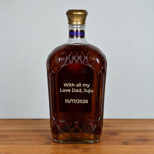 A personalized bottle of crown royal whiskey with a gold cap and a custom-engraved label reading 'With all my Love Dad, Juju 10/17/2026', displayed on a wooden surface against a neutral gray background.