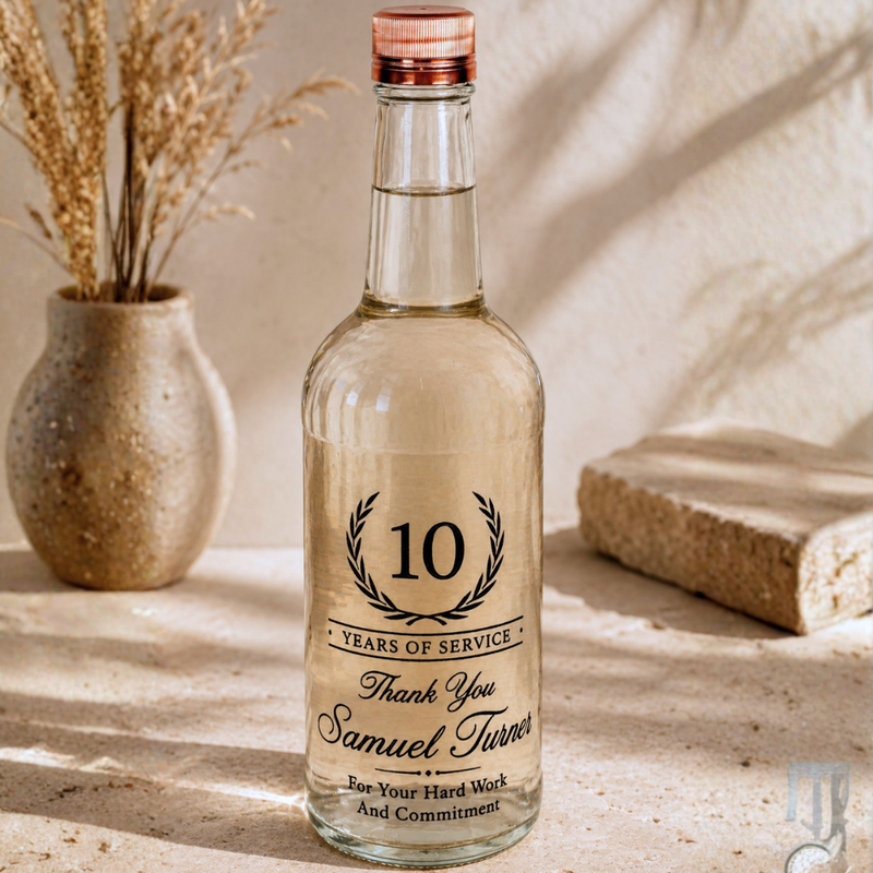 A clear glass bottle with a metallic rose gold cap and a black printed '10 Years of Service' thank you label, styled on a stone surface next to a ceramic vase with dried grass.
