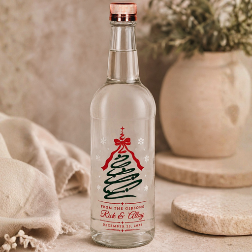 A clear glass bottle with a personalized Christmas tree label, standing on a stone coaster with a neutral beige background and soft-focus decorative props.